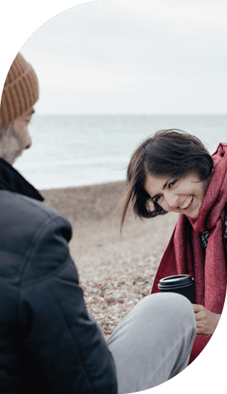 Frau mit rotem Schal sitzt am Strand und schaut lachend zur Seite