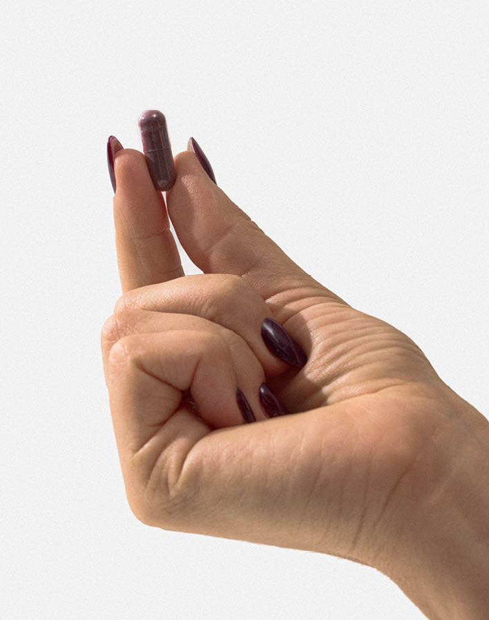 Hand mit dunkellackierten Fingernägeln hält eine einzelne Kapsel zwischen Daumen und Zeigefinger