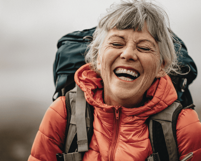 Lächelnde ältere Frau mit grauem Haar und roter Outdoor-Jacke mit Rucksack